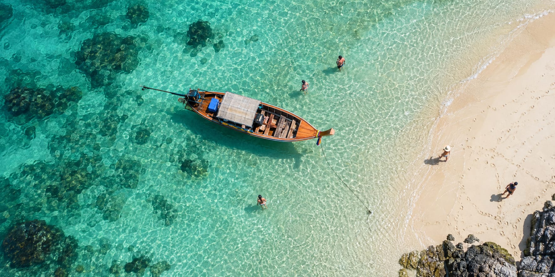 Una barca a coda lunga ormeggiata in acqua turchese trasparente accanto a una spiaggia bianca, vista dall’alto, con qualcuno che cammina sulla sabbia.